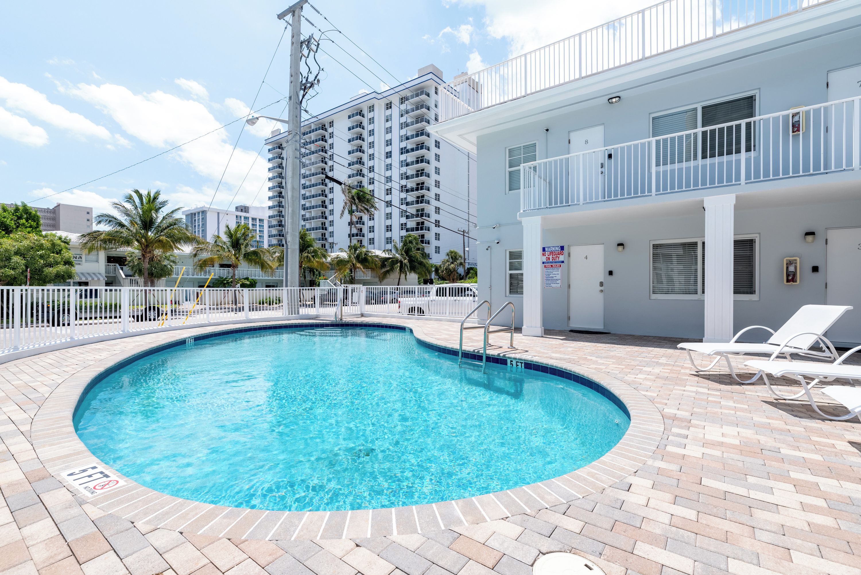 A swimming pool with a brick deck, white lounge chairs, and a white building with balconies.
