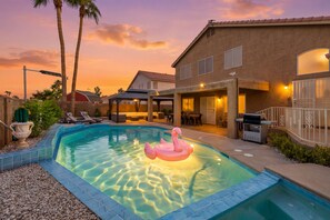Ein Wohnpool mit einem Flamingo-Schwimmdings, umgeben von einer Terrasse mit Gartenmöbeln, einer überdachten Sitzgruppe und einem Haus mit Ziegeldach.