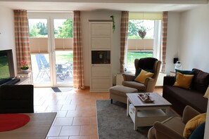 A living room with a sofa, armchair, coffee table, and a television. There is a sliding glass door leading to an outdoor area with a patio table and chairs. - Meerluft Boltenhagen Vacation Rental - Sünnslag Complex (Boltenhagen)