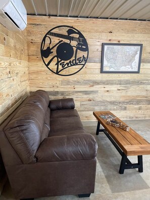 A room with a brown leather sofa, a wooden coffee table, and a wall with a Fender logo and a framed map. - The Rock-N-Roll Cabin with Meramec River Access (Liberty Township)