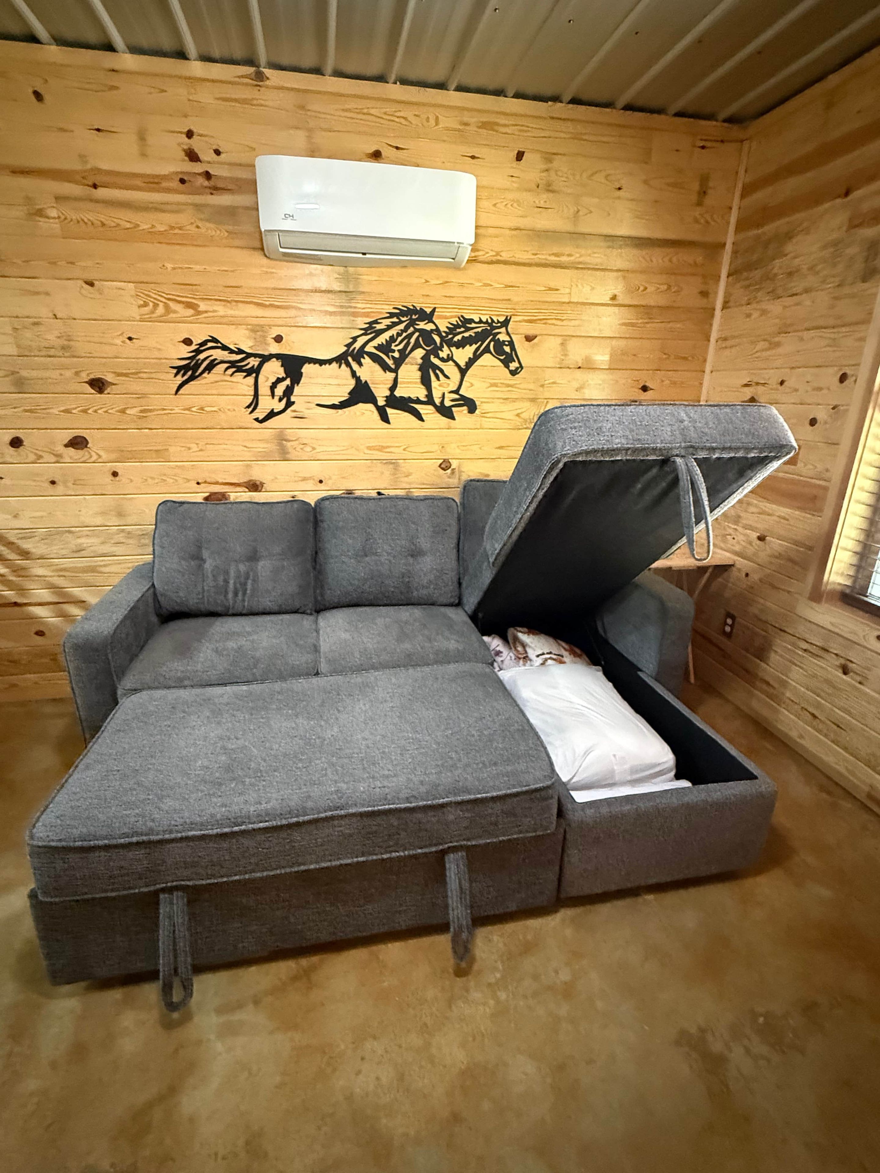 A grey sofa with a storage compartment, a wooden wall with a horse decal, and an air conditioning unit.