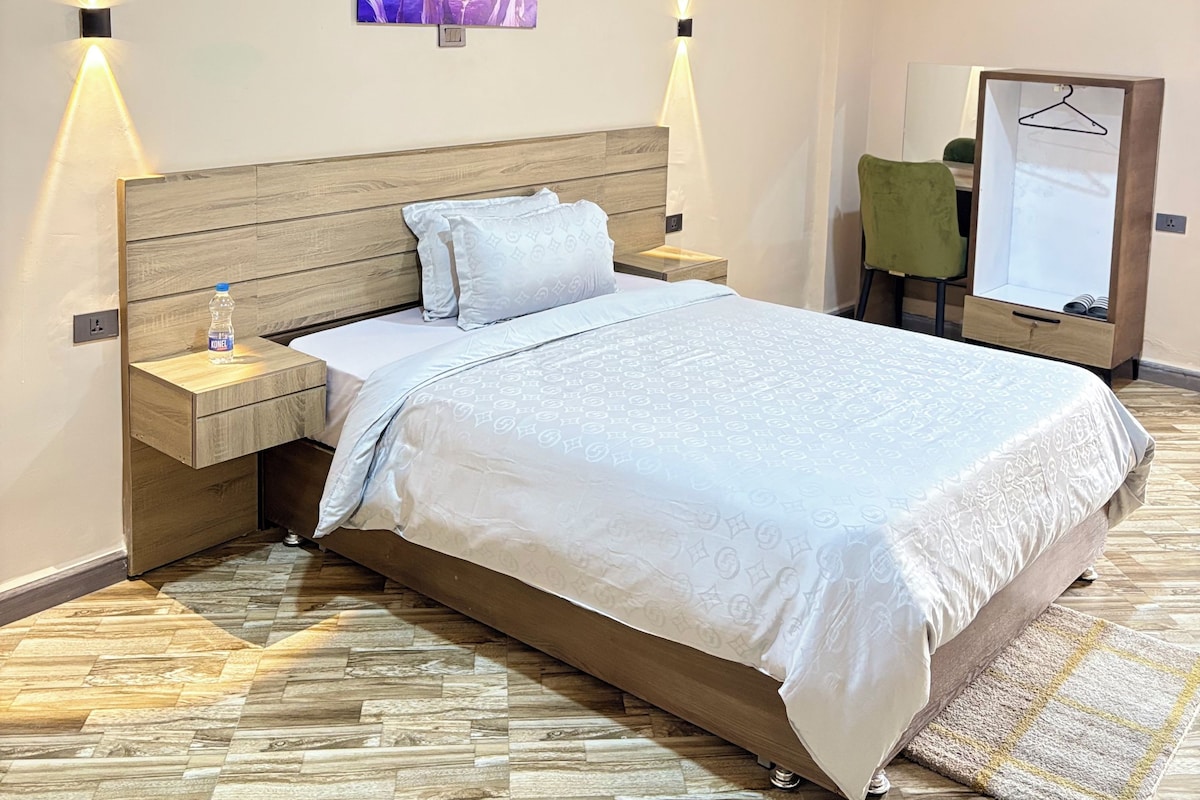 A hotel room with a large bed, wooden headboard, two bedside tables, a water bottle, a painting, and a mirror on the wall.