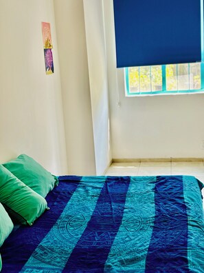 Ein Zimmer mit einem Bett, grünen Kissen, einer Bettdecke in Blau und Schwarz gebürstet und einem Fenster mit blauer Jalousie sowie einer kleinen Wanddekoration.