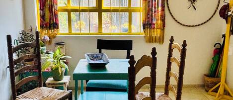 Una habitación con una mesa y cuatro sillas, una ventana con cortinas, un atrapasueños en la pared y una planta en maceta sobre la mesa.