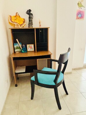 Ein Zimmer mit einem Holztisch, einem Stuhl mit blauer Sitzkissen sowie einem Regal mit verschiedenen Gegenständen, darunter eine Statue, ein gerahmtes Bild und eine kleine Vase.