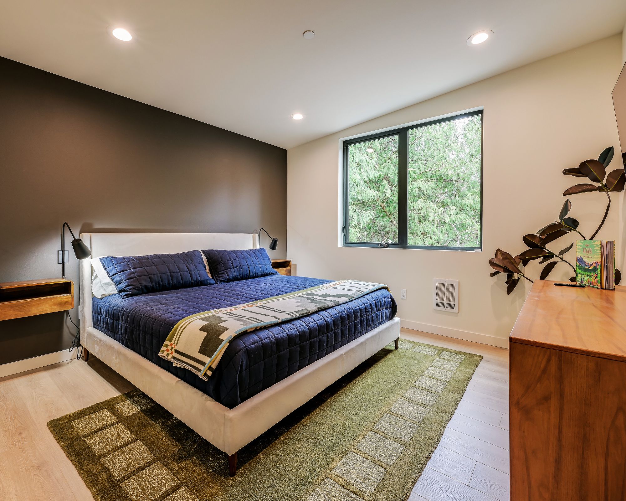 A modern bedroom with a large bed, a window with a view of trees, a bedside table, and a nightstand.