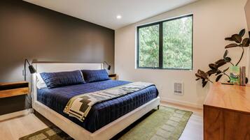 A modern bedroom with a large bed, a window with a view of trees, a bedside table, and a nightstand.