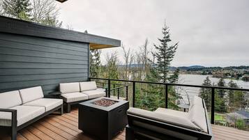 A deck with a sofa, chairs, and a fire pit, overlooking a lake and forest.