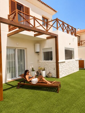 A person relaxing on a wooden lounge chair in a covered outdoor area with artificial grass, a stone wall, and potted plants.