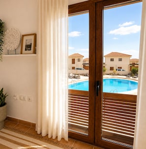 A room with a view of a swimming pool, a wooden door, a white curtain, a framed picture, and a potted plant.