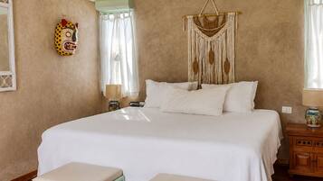 A bedroom with a large bed, two bedside tables, a hanging macrame wall decoration, and a colorful mask on the wall.
