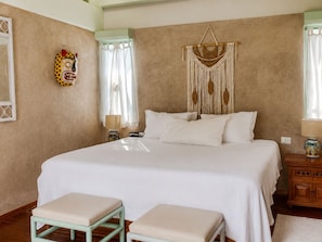 Un dormitorio con una cama grande, dos mesitas de noche, un adorno de macramé en la pared y una máscara colorida también en la pared.