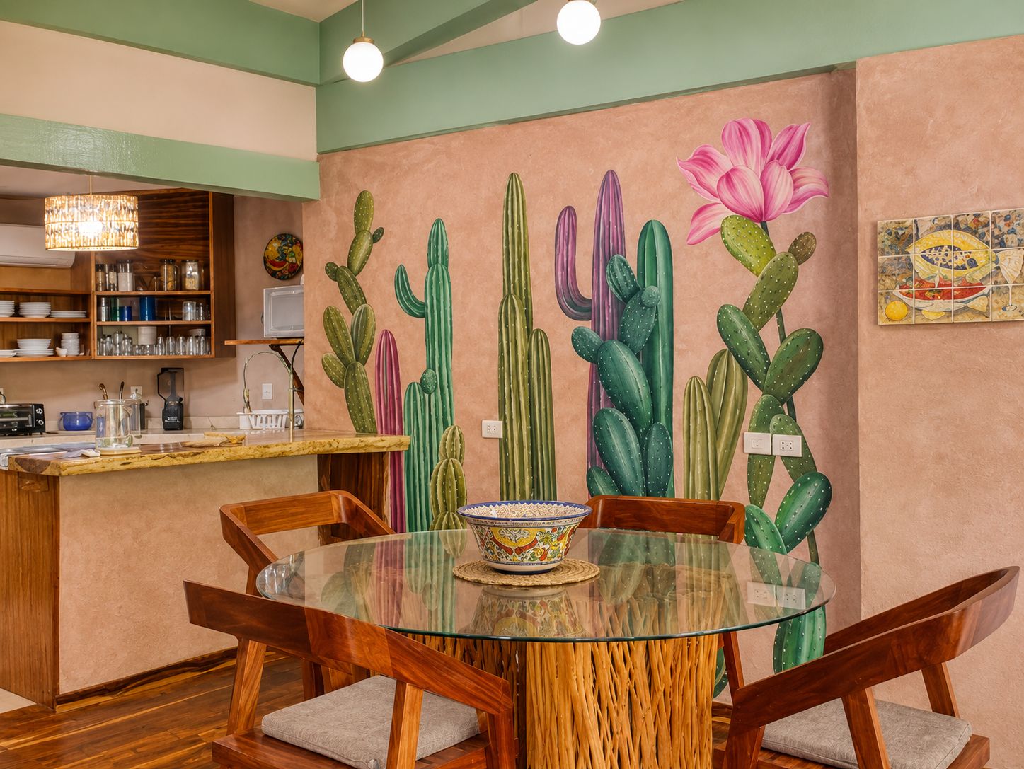 A dining area with a glass table surrounded by wooden chairs, a cactus mural on the wall, and a kitchen area with shelves and appliances in the background.