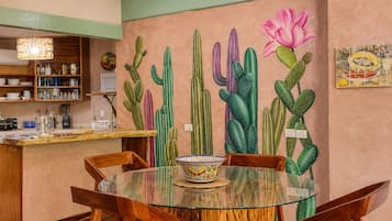 A dining area with a glass table surrounded by wooden chairs, a cactus mural on the wall, and a kitchen area with shelves and appliances in the background.