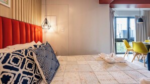 Une chambre d’hôtel avec un lit, une tête de lit rouge, un luminaire suspendu, une fenêtre avec des rideaux et un coin repas avec des chaises jaunes.