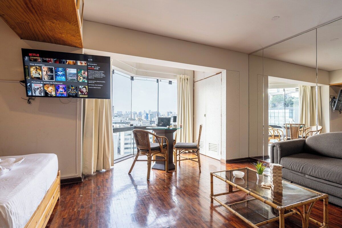 Habitación de hotel con cama, sofá, zona de comedor con sillas, televisor montado en la pared y un amplio ventanal con vista a la ciudad.