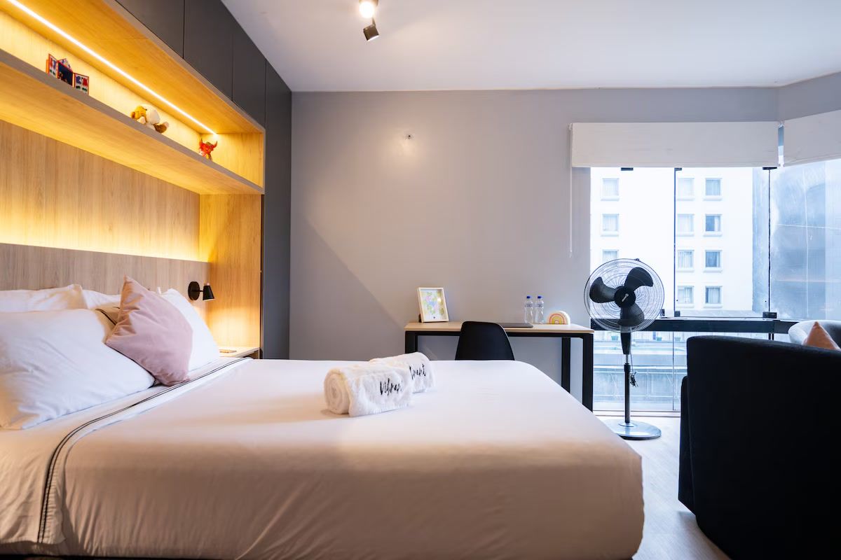 Une chambre d’hôtel équipée d’un lit, d’une table de chevet, d’un bureau, d’une chaise et d’une fenêtre donnant sur des immeubles.