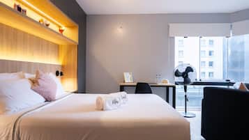 Une chambre d’hôtel équipée d’un lit, d’une table de chevet, d’un bureau, d’une chaise et d’une fenêtre donnant sur des immeubles.