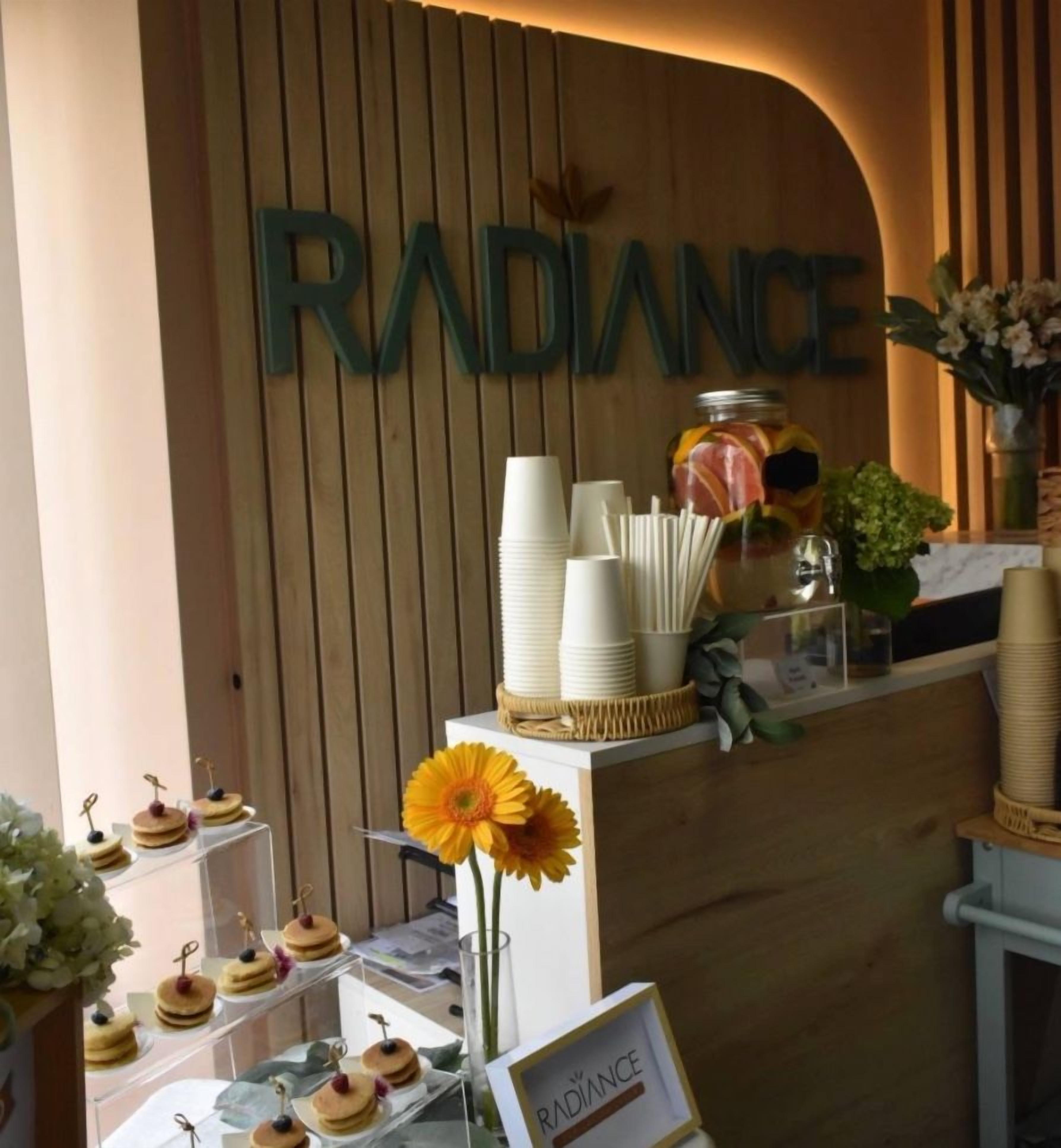 Un área de recepción con un mostrador de madera, un cartel que dice 'RADIANCE', y diversos objetos como vasos de papel, un jarrón con flores y un tarro de aperitivos.