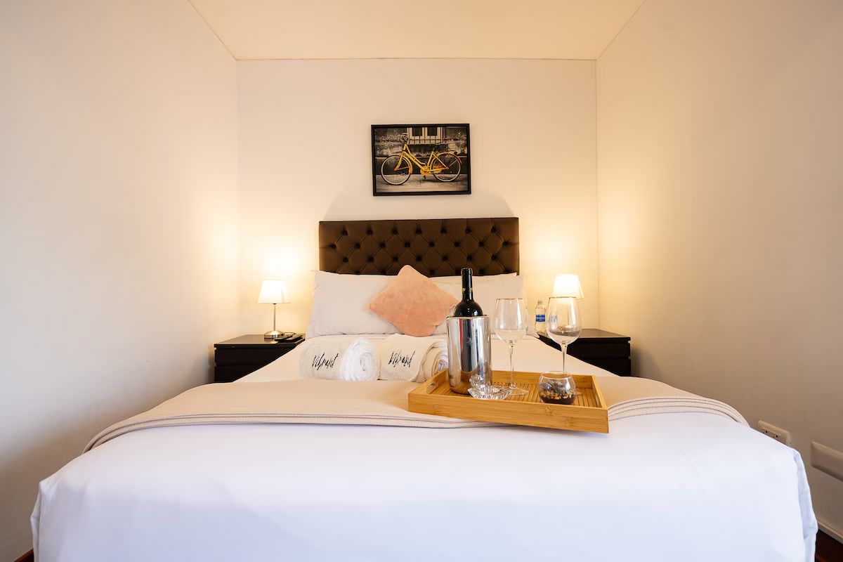 Ein Hotelzimmer mit einem Bett, einem Kopfteil, einem Nachttisch mit Lampe, einem Tablett mit Flasche und Gläsern sowie einem gerahmten Bild an der Wand.