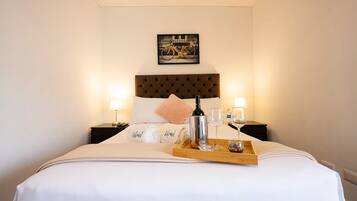 Ein Hotelzimmer mit einem Bett, einem Kopfteil, einem Nachttisch mit Lampe, einem Tablett mit Flasche und Gläsern sowie einem gerahmten Bild an der Wand.