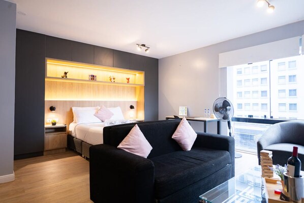 A modern hotel room with a bed, a black sofa, a bedside table with a lamp, a fan, and a window with a view of buildings. - Chateau Studios by Vibrant (Miraflores)