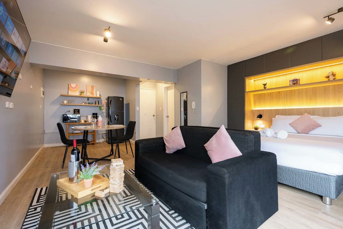 A modern living room with a black sofa, a dining area with a table and chairs, a bed with a headboard, and a wall-mounted shelf with decorative items.