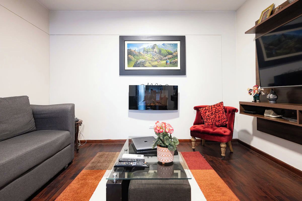 Una sala de estar con un sofá gris, un sillón rojo, una mesa de centro con controles remotos, una computadora portátil y un cuadro de un paisaje en la pared.