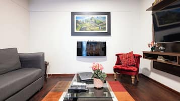 Een woonkamer met een grijze bank, een rode fauteuil, een salontafel met afstandsbedieningen, een laptop, en een lijst met een landschapsafbeelding aan de muur.
