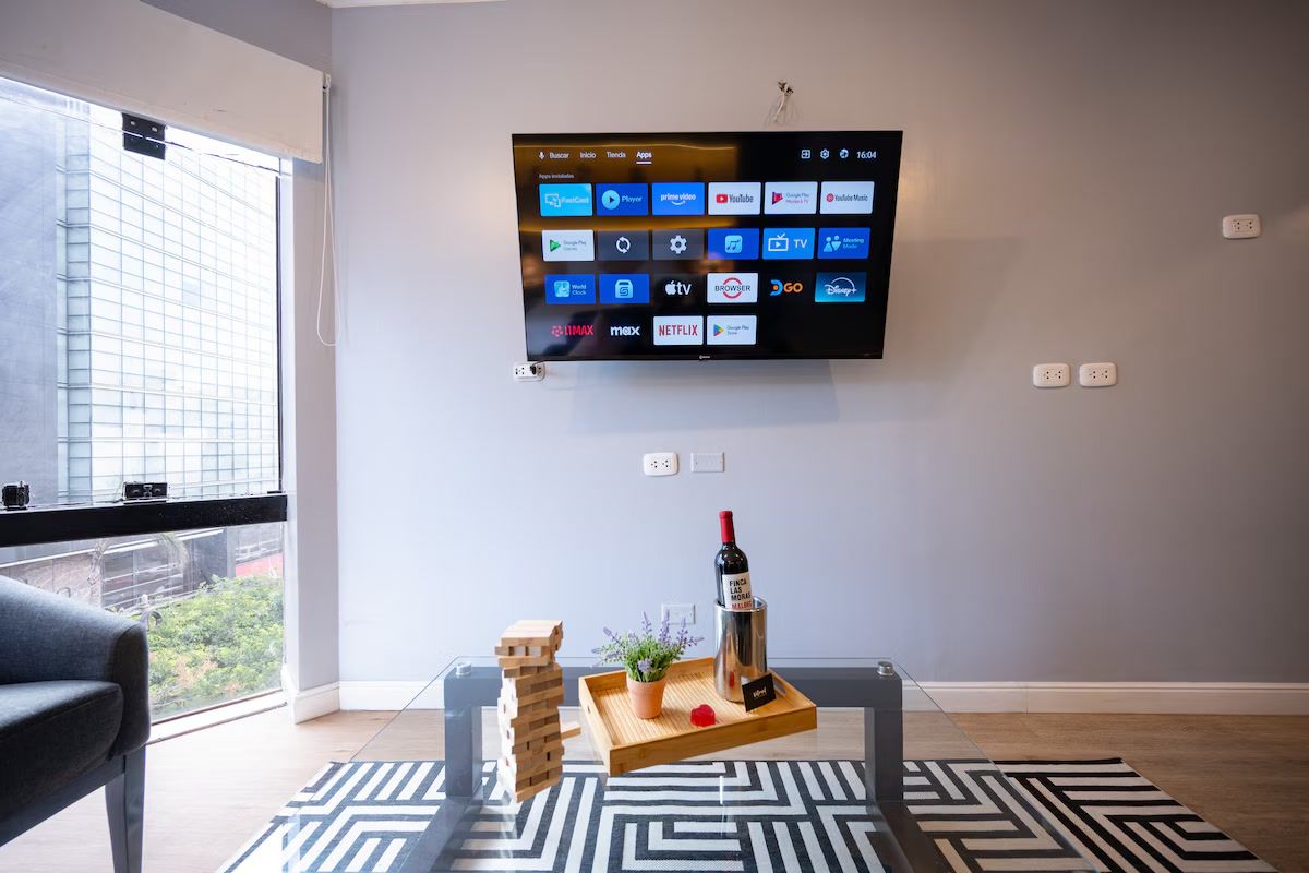 Una sala de estar moderna con un televisor de pantalla plana montado en la pared que muestra varias aplicaciones de transmisión, una mesa de centro de vidrio con una botella de vino, una bandeja con una planta y un objeto rojo, y una ventana con vista a un edificio.