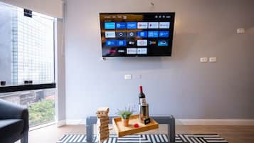 Een moderne woonkamer met een flatscreen televisie aan de muur waarop verschillende streamingdiensten zichtbaar zijn, een glazen salontafel met een fles wijn, een dienblad met een plant en een rood object, en een raam met uitzicht op een gebouw.