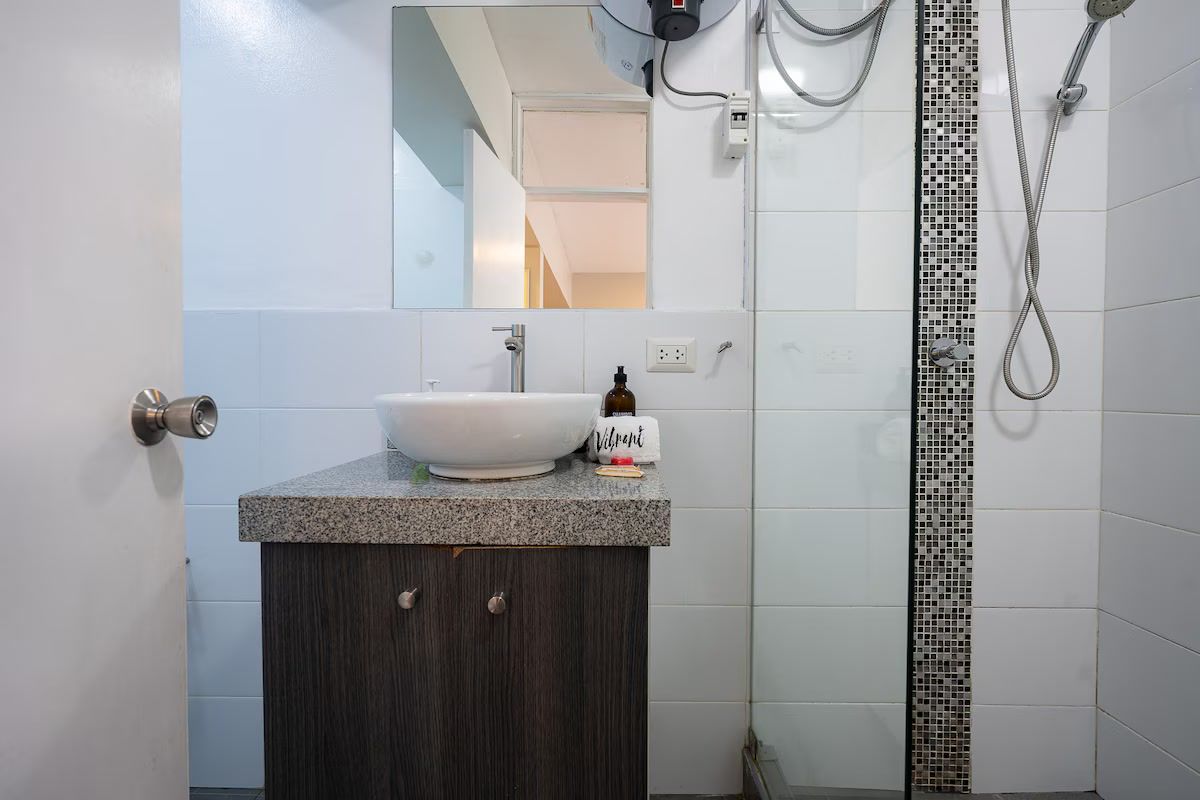 Un baño con un lavamanos blanco, un mueble de madera oscura y una ducha con pared de azulejos.
