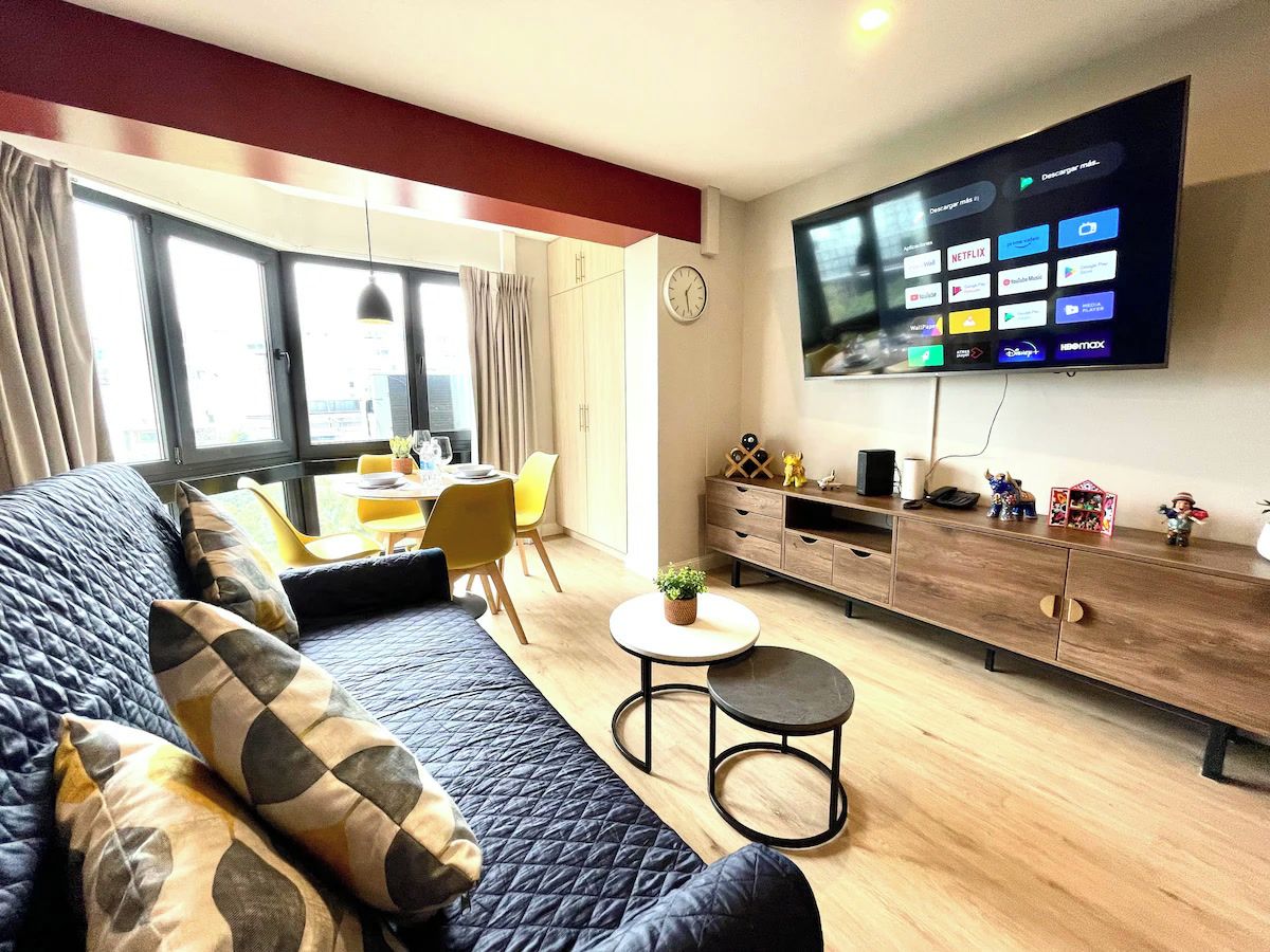A modern living room with a large flat-screen TV, a sofa, a coffee table, and a dining area with yellow chairs.