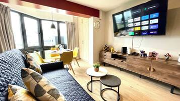 A modern living room with a large flat-screen TV, a sofa, a coffee table, and a dining area with yellow chairs.