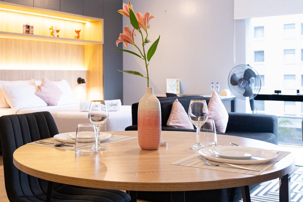 Un comedor con una mesa redonda preparada para dos personas, un jarrón con flores y un sofá al fondo.