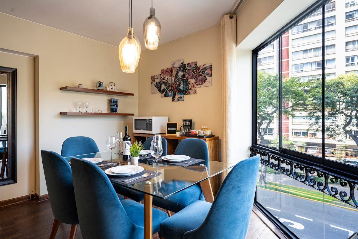 A dining area with a glass table, blue chairs, a kitchen in the background, and a city view through large windows.