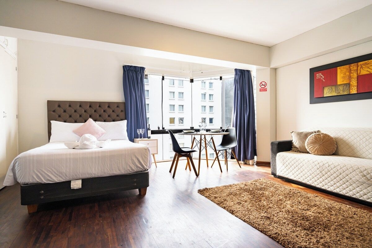 A hotel room with a bed, a sofa, a dining table with chairs, and a view of buildings through large windows.