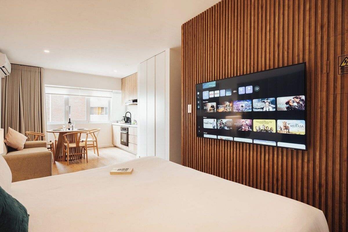 A modern hotel room with a large flat-screen TV mounted on a wooden wall, a bed, a dining area with a table and chairs, and a kitchenette in the background.