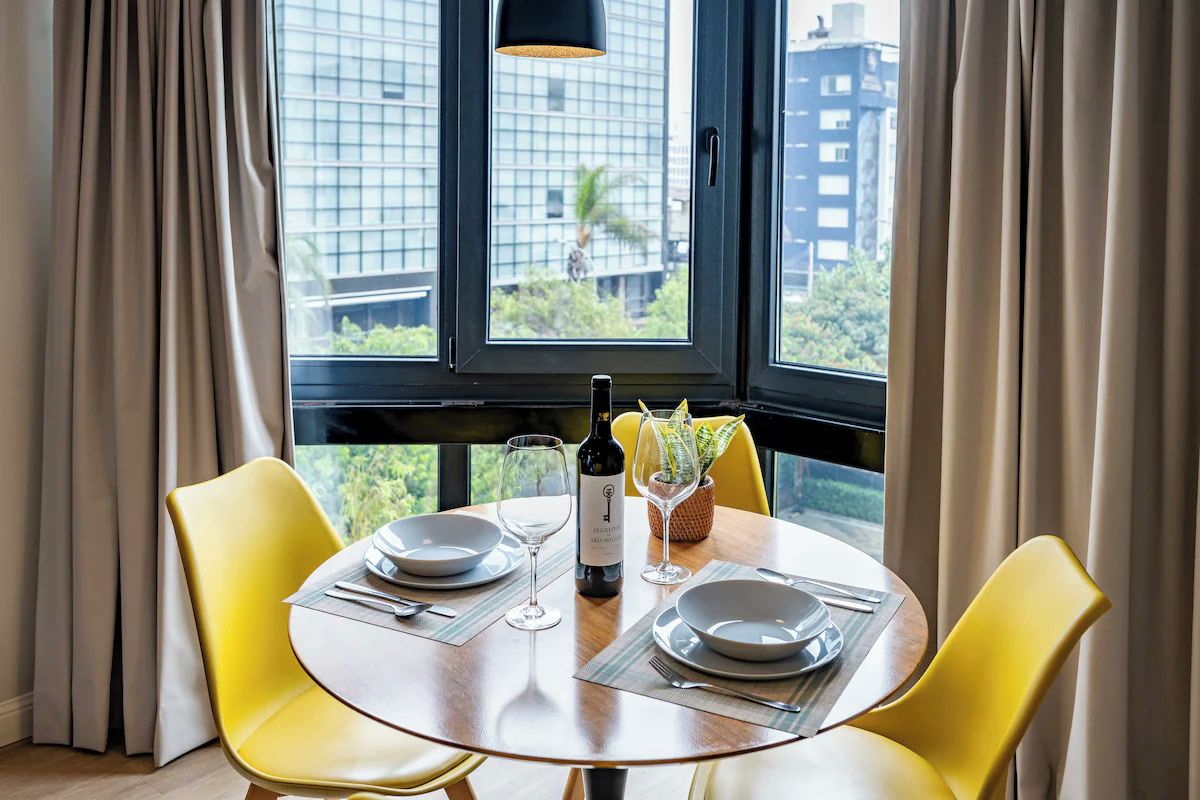 Une table à manger avec des chaises jaunes, une bouteille de vin et un verre, le tout placé devant une fenêtre donnant sur un paysage urbain.