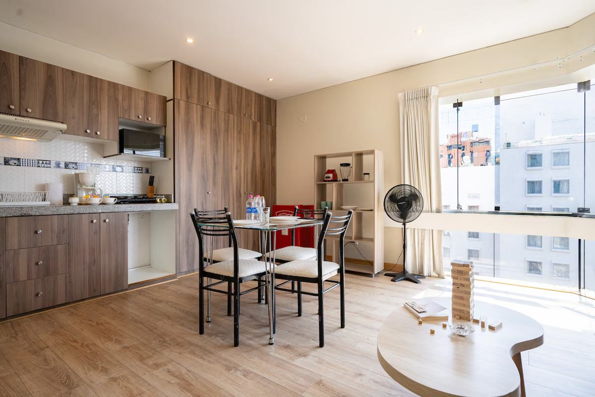 Una cocina moderna con armarios de madera, un comedor con sillas, un ventilador y vistas a edificios a través de amplios ventanales.