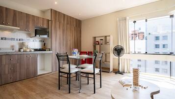 Una cocina moderna con armarios de madera, un comedor con sillas, un ventilador y vistas a edificios a través de amplios ventanales.