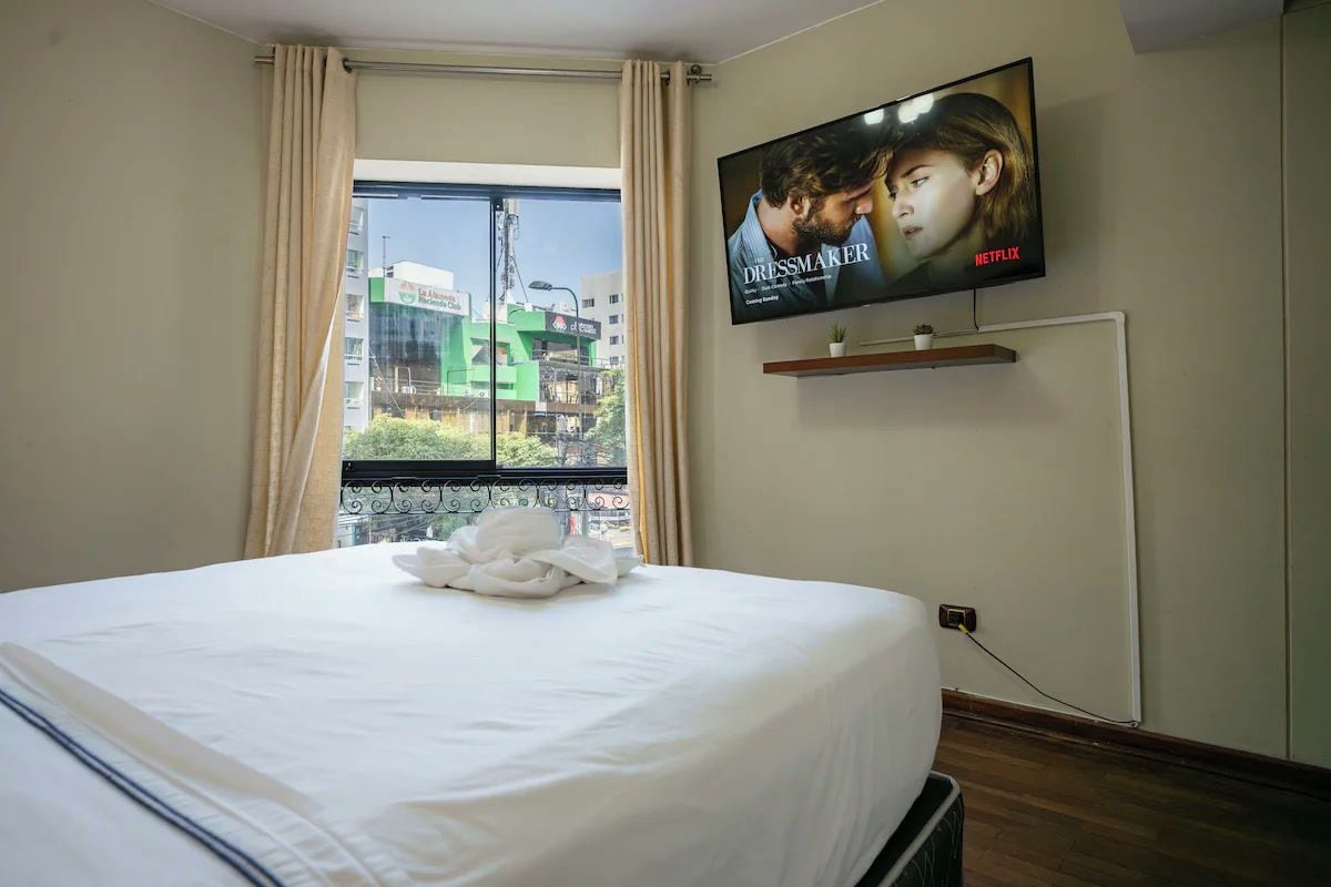A hotel room with a bed, a TV mounted on the wall, a window with curtains, and a view of the cityscape.