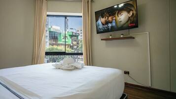 A hotel room with a bed, a TV mounted on the wall, a window with curtains, and a view of the cityscape.