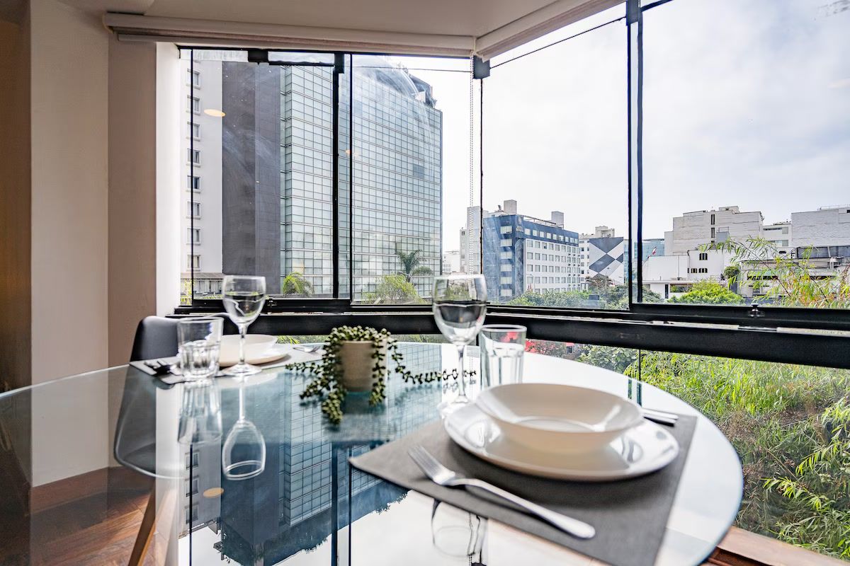 Mesa de comedor servida con platos, cubiertos y copas de vino, ubicada cerca de un ventanal con vista a la ciudad.