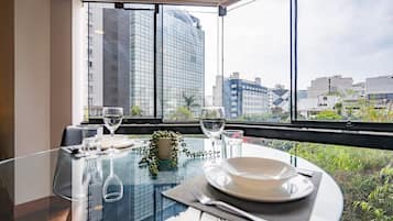 Mesa de comedor servida con platos, cubiertos y copas de vino, ubicada cerca de un ventanal con vista a la ciudad.