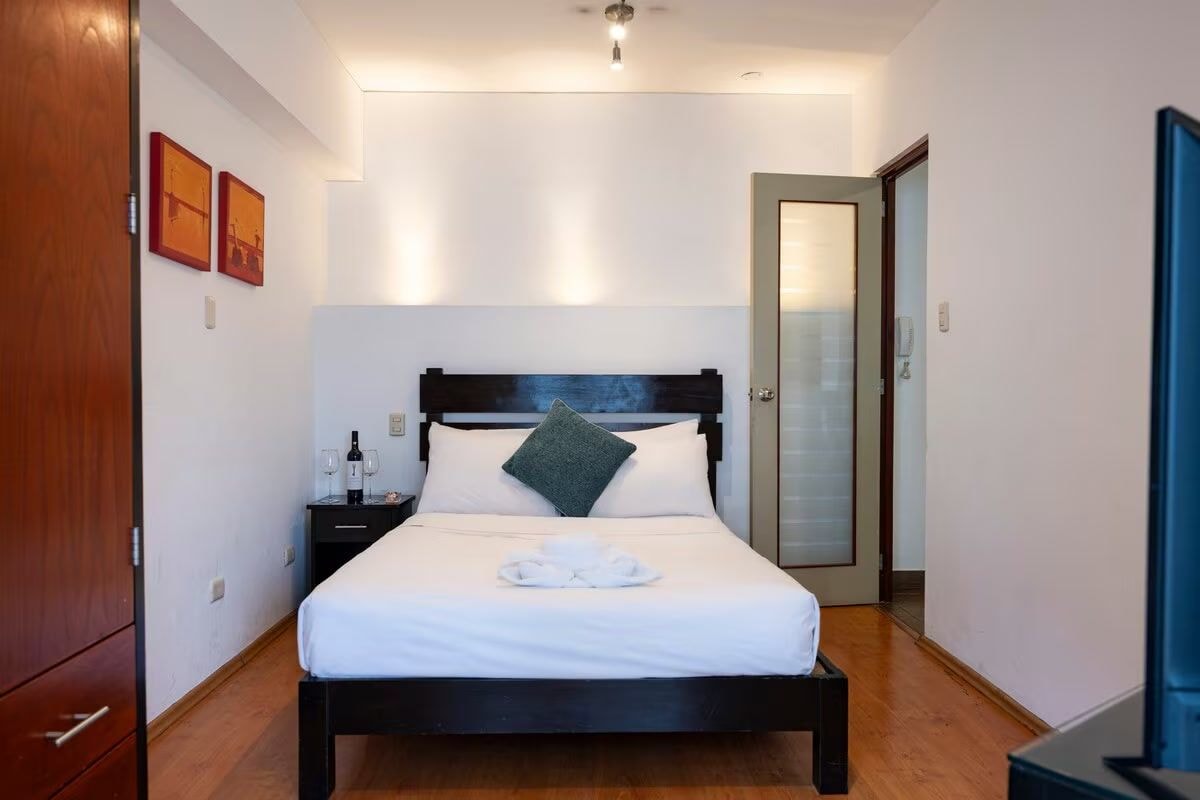A hotel room with a bed, a nightstand, a bottle of wine, and a towel on the bed.
