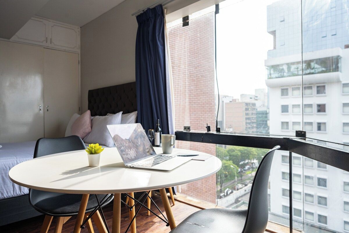 Une chambre avec un lit, un ordinateur portable posé sur une table ronde, une plante en pot et une vue sur des immeubles par la fenêtre.