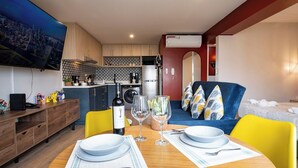 Un espace de vie compact comprenant un coin repas, une kitchenette et un téléviseur diffusant des images d’un paysage urbain.