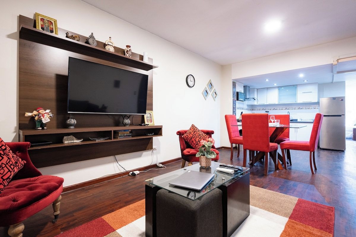 A modern living room with a flat-screen TV, red chairs, a glass coffee table, and a kitchen in the background.