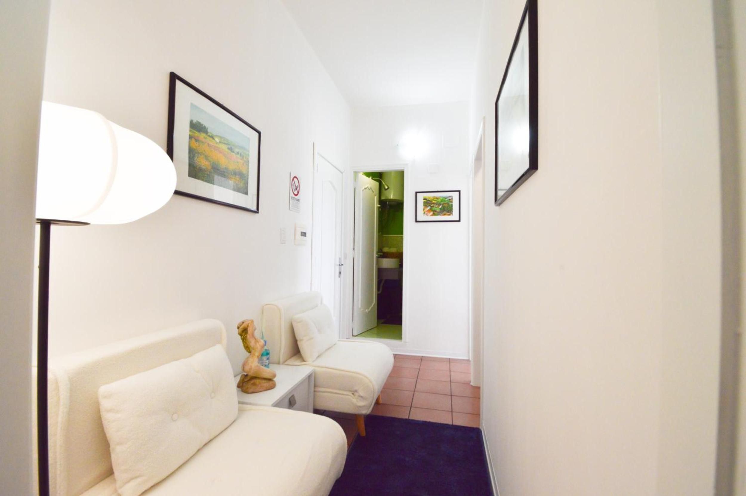 Un pasillo con un sofá blanco, una mesita y una lámpara. Hay cuadros en las paredes y una puerta que da a otra habitación.
